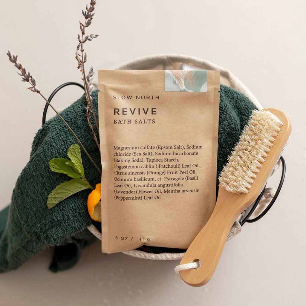 Enjoy Slow North's Revive bath salts for the ultimate rejuvenation experience. Revive is pictured with a scrubbing brush, a forest green cloth, mint leaves, and the subtle peel of an orange. 